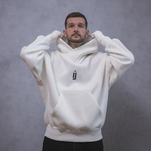 May include: Cream-colored hoodie with a large front pocket and a hood. The hoodie features a small black logo on the chest. The person in the image is wearing the hoodie and pulling up the hood. The background is a solid gray color.