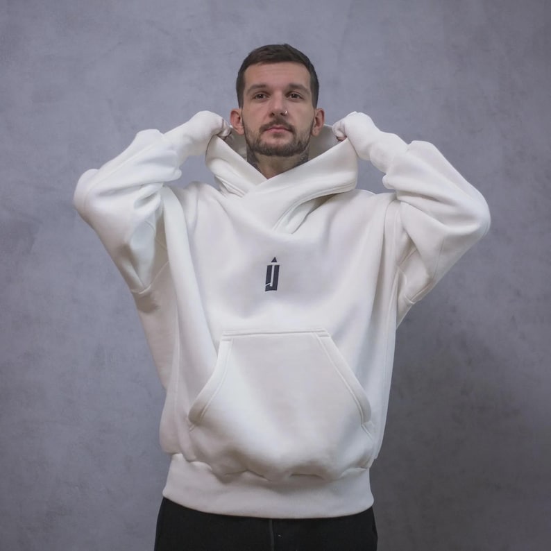 May include: Cream-colored hoodie with a large front pocket and a hood. The hoodie features a small black logo on the chest. The person in the image is wearing the hoodie and pulling up the hood. The background is a solid gray color.
