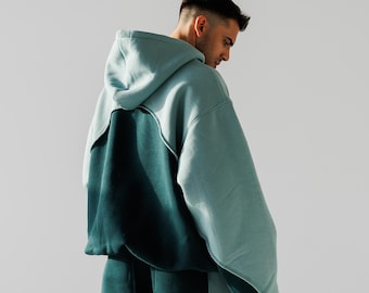 May include: A hooded sweatshirt with a color block design in shades of green and blue. The sweatshirt has a relaxed fit and a large hood.