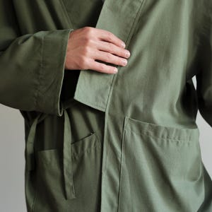 May include: An olive green robe with a wrap-around design. It has a shawl collar, two front pockets, and a matching belt. The sleeves are long, and the fabric looks like a lightweight cotton or linen blend. The robe is designed for comfort and relaxation.