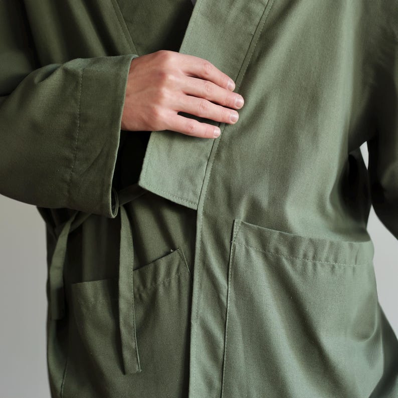 May include: An olive green robe with a wrap-around design. It has a shawl collar, two front pockets, and a matching belt. The sleeves are long, and the fabric looks like a lightweight cotton or linen blend. The robe is designed for comfort and relaxation.