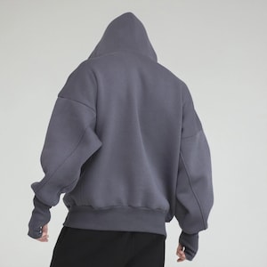 May include: A gray hooded sweatshirt with a relaxed fit. The sweatshirt features a large hood, long sleeves with thumbhole cuffs, and a ribbed hem. The back view shows the garment's oversized silhouette and solid color.