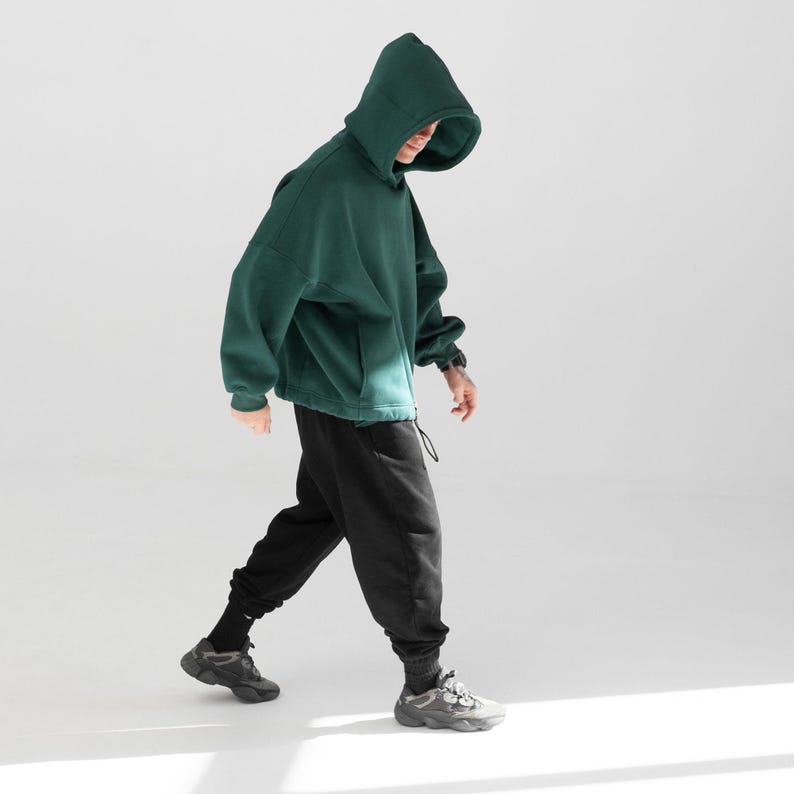 May include: A person wearing a dark green hooded sweatshirt with a drawstring and black sweatpants. The person is wearing gray sneakers and black socks. The outfit is casual and comfortable.
