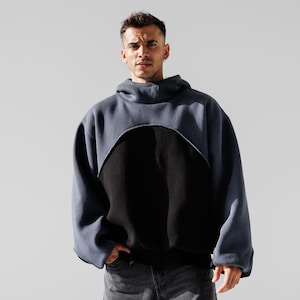 May include: A two-tone hoodie in a modern style. The upper section is a muted blue-gray, with a curved cutout revealing a black panel. The hoodie has a high collar and long sleeves. The model is wearing gray jeans.