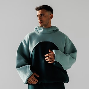May include: A two-tone hoodie in teal and dark teal. The teal portion covers the hood and upper body, with a large circular cutout revealing the dark teal lower section. The sleeves are long and loose, with wide cuffs.