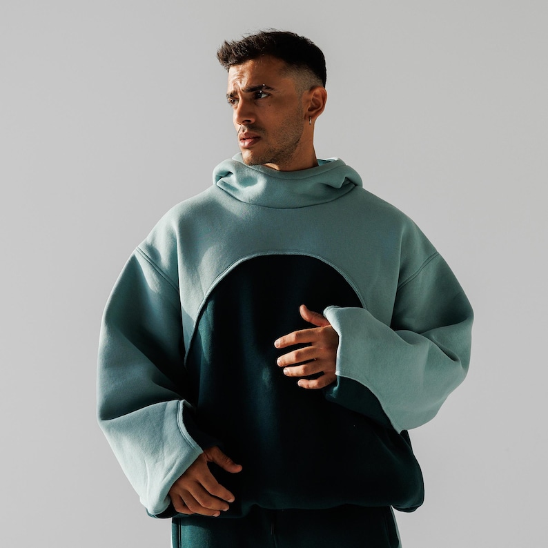 May include: A two-tone hoodie in teal and dark teal. The teal portion covers the hood and upper body, with a large circular cutout revealing the dark teal lower section. The sleeves are long and loose, with wide cuffs.