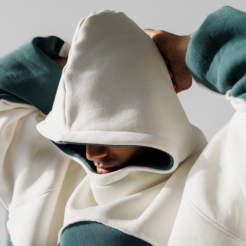 May include: A close-up of a person wearing a white hoodie with a teal-colored interior. The hoodie is made of a soft material and has a unique design. The person's face is partially visible, with their lips and nose showing. The background is a plain white.