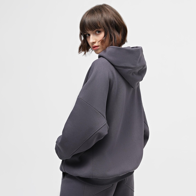 May include: A charcoal gray hoodie with a large hood and a relaxed fit. The hoodie has a simple, minimalist design, with seams visible on the sleeves and shoulders. The model is wearing a matching pair of sweatpants.