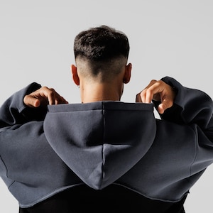 May include: A dark gray cropped hoodie with a hood, viewed from the back. The person is pulling up the hood. The hoodie is over a black shirt. The background is a plain white.