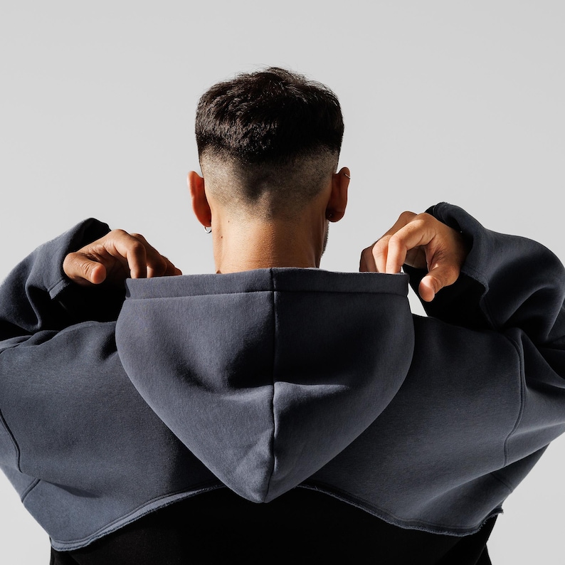 May include: A dark gray cropped hoodie with a hood, viewed from the back. The person is pulling up the hood. The hoodie is over a black shirt. The background is a plain white.