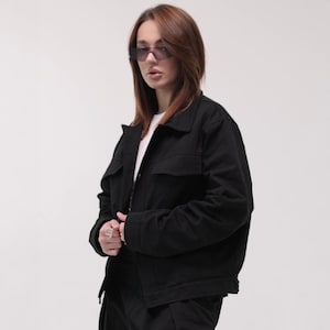 Könnte beinhalten: Schwarze Jacke mit zwei Brusttaschen und einem Kragen. Die Jacke wird über einem weißen Hemd getragen. Die Person auf dem Bild trägt auch dunkle Sonnenbrillen.