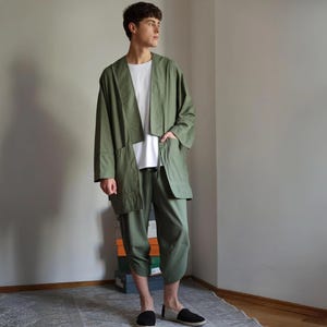 May include: An olive green outfit comprising a jacket, a white t-shirt, and cropped trousers. The jacket features an open design with large pockets. The trousers are a matching olive green. The outfit is paired with black and grey slip-on shoes.