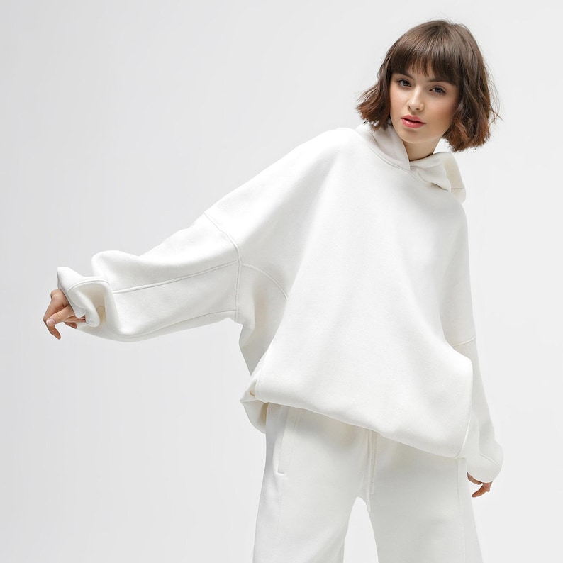 May include: A white hoodie with a matching pair of sweatpants. The hoodie has a drawstring hood and a kangaroo pocket. The outfit is made of a soft, comfortable material. The model is wearing the outfit.