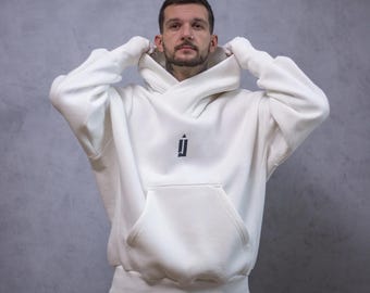 Może przedstawiać: Kremowa bluza z kapturem z dużą przednią kieszenią i kapturem. Bluza ma małe czarne logo na piersi. Osoba na zdjęciu ma na sobie bluzę z kapturem i podnosi kaptur.