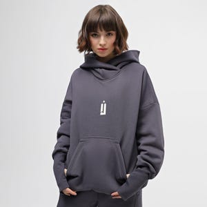 May include: A dark gray hoodie with a white logo on the chest. The hoodie has a large front pocket and a hood. The model is wearing the hoodie and is standing in front of a white background. The hoodie is made of a soft material.