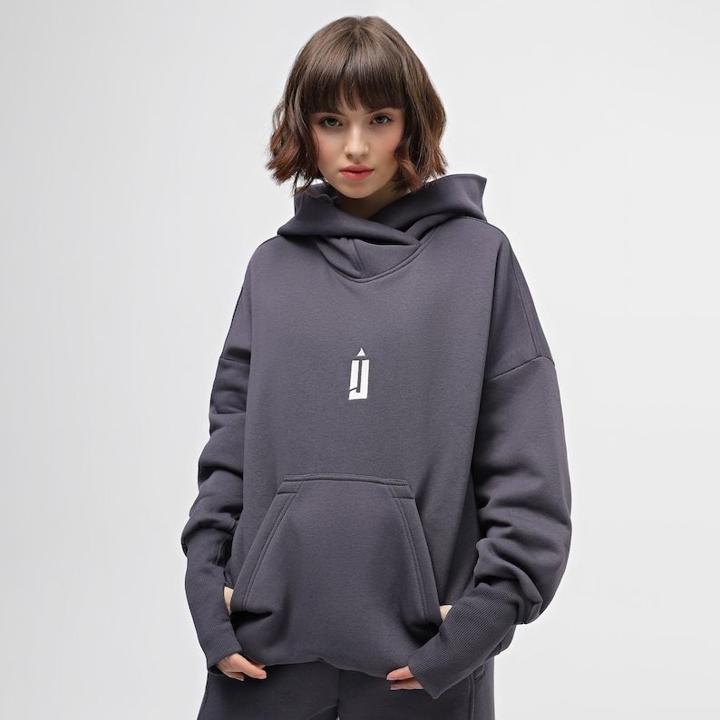 May include: A dark gray hoodie with a white logo on the chest. The hoodie has a large front pocket and a hood. The model is wearing the hoodie and is standing in front of a white background. The hoodie is made of a soft material.