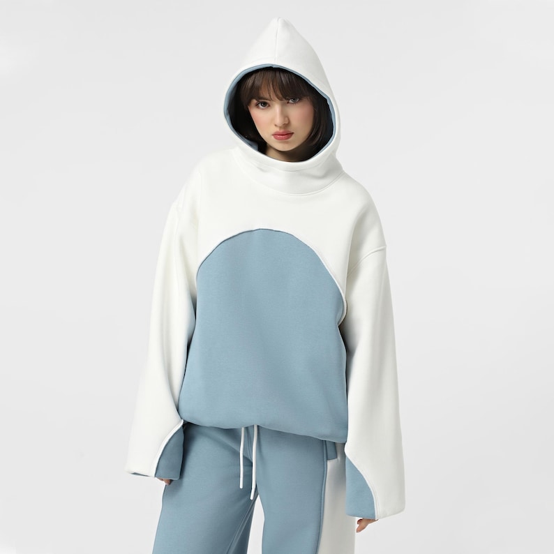 May include: A two-tone hooded sweatshirt in white and light blue. The sweatshirt features a unique design with a high collar and a drawstring at the waist. The sleeves are long and wide, and the hood is lined with a contrasting blue.