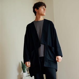 May include: A black kimono-style jacket with large front pockets. The jacket is worn over a grey t-shirt and black trousers. The jacket has a relaxed fit and a draped, open front design. The outfit is simple and modern.