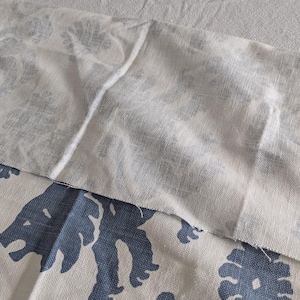 May include: A white fabric with a subtle pattern is layered on top of a white fabric with a blue pattern of stylized lions. The fabric is textured and has a slightly rough surface.