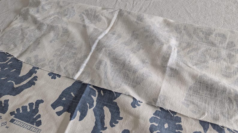 May include: A white fabric with a subtle pattern is layered on top of a white fabric with a blue pattern of stylized lions. The fabric is textured and has a slightly rough surface.