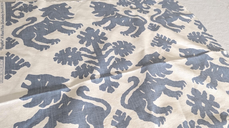 May include: A white fabric with a repeating pattern of blue stylized lions and floral designs. The fabric has a subtle texture.