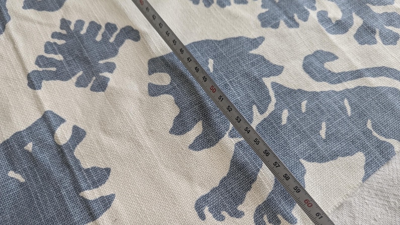 May include: A white fabric with a blue pattern of stylized animal shapes. A metal ruler is placed diagonally across the fabric, with the numbers 49 through 61 visible.