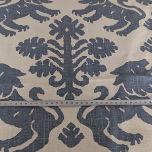 May include: A white fabric with a blue repeating pattern of stylized lions and floral designs. The fabric is measured with a tape measure.