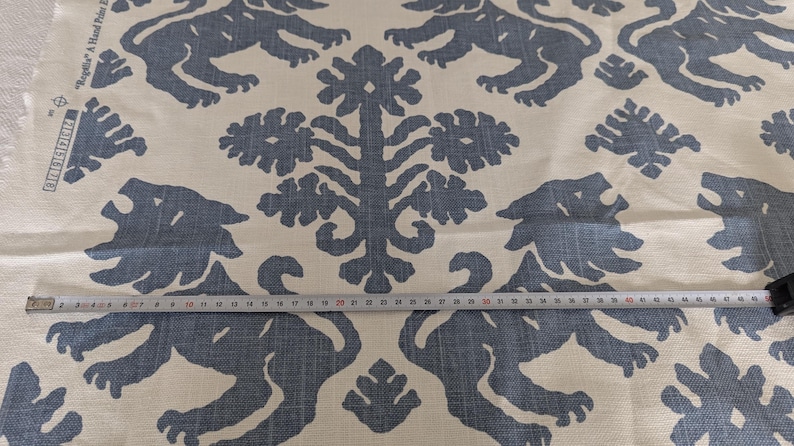 May include: A white fabric with a blue repeating pattern of stylized lions and floral designs. The fabric is measured with a tape measure.