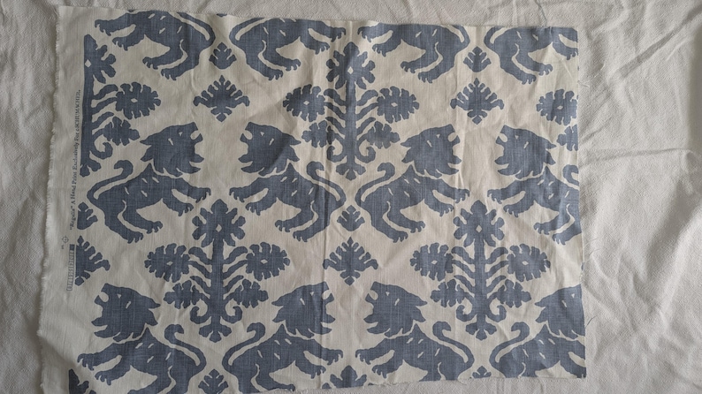 May include: A white fabric with a repeating pattern of blue stylized lions and floral designs. The fabric has a slightly textured surface.