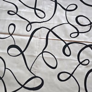 May include: Cream-colored fabric with a repeating pattern of black, flowing, abstract lines. The lines twist and turn, creating loops and curves across the surface. The fabric appears to be folded in places.