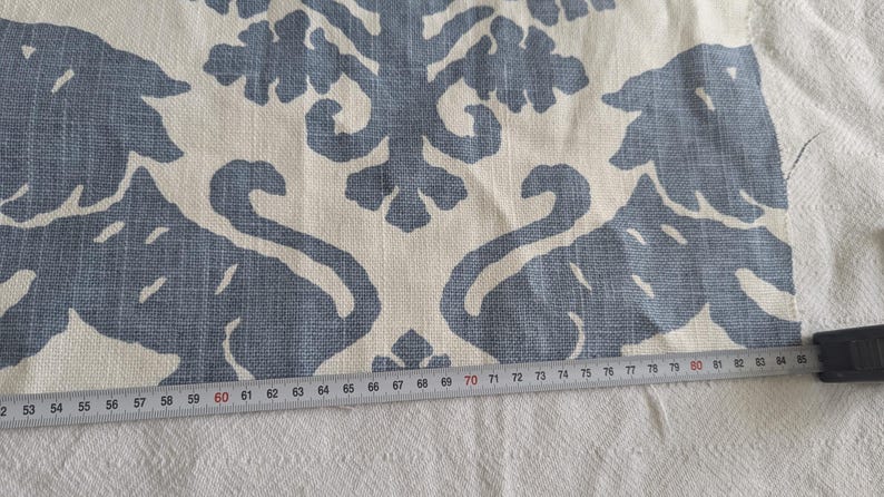 May include: A close-up of a white fabric with a blue patterned design. The pattern features stylized swans and leaves. A ruler is placed on the fabric, showing measurements in inches.