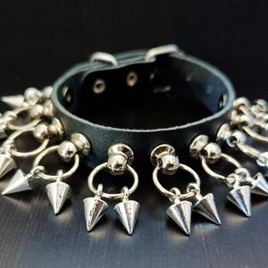 May include: Black leather choker with silver-colored cone spikes and metal ring accents. The choker has a buckle closure and multiple rows of spikes hanging from the band. The jewelry is set against a dark background.