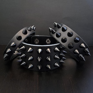 Peut inclure: Trois bracelets de poignet en cuir noir avec des clous coniques argentés. Les bracelets ont des largeurs variables et des fermetures à pression. Le fond est de couleur sombre et unie.