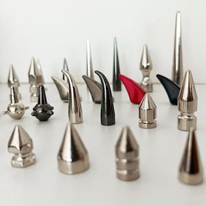 May include: A selection of metal decorative spikes and studs in silver, black, and red. The collection features cone-shaped, curved, and cylindrical designs, with various sizes and finishes. The objects are arranged on a white surface.