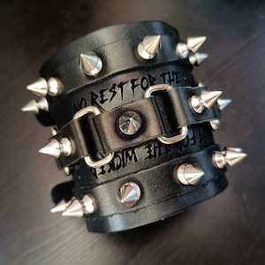 May include: Black leather wristband with silver metal spikes and the text "No Rest For The Wicked" engraved on it.