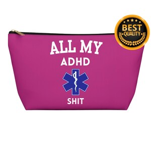 以下が含まれることがあります： 白い文字で「ALL MY ADHD SHIT」と書かれたピンクのジッパー付きメイクアップバッグ。バッグの中央には青と白の医療シンボルがあります。