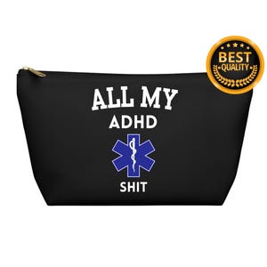 以下が含まれることがあります： 白い文字で「ALL MY ADHD SHIT」と書かれた黒いキャンバス製のジッパー付きポーチ。テキストの下に青と白の医療シンボルが中央に配置されています。
