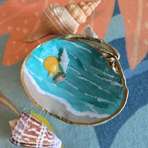 May include: A painted seashell dish with a gold rim, featuring a miniature beach scene with turquoise water and white waves. Small seashells are placed inside. Other seashells are scattered around the dish.