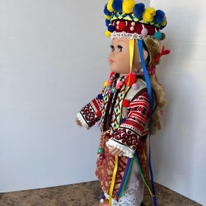 May include: A porcelain doll wearing a traditional folk costume with a colorful embroidered vest, a red and white skirt, and a crown with yellow and blue pom-poms.