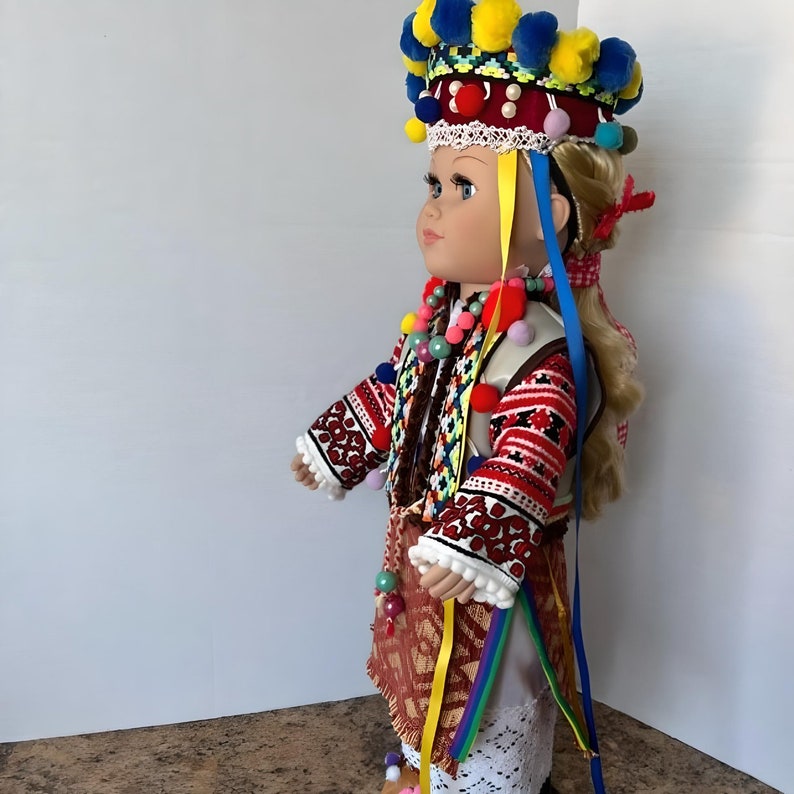 May include: A porcelain doll wearing a traditional folk costume with a colorful embroidered vest, a red and white skirt, and a crown with yellow and blue pom-poms.