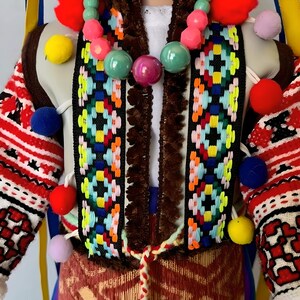 May include: A colorful folk costume with a red, white, and blue embroidered pattern. The costume features a brown fur trim, a multi-colored beaded necklace, and pom-poms in various colors.