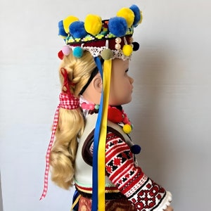 May include: A doll wearing a traditional folk costume with a colorful headpiece featuring pom-poms and ribbons. The doll has blonde hair and is wearing a red and white embroidered shirt with a white vest.