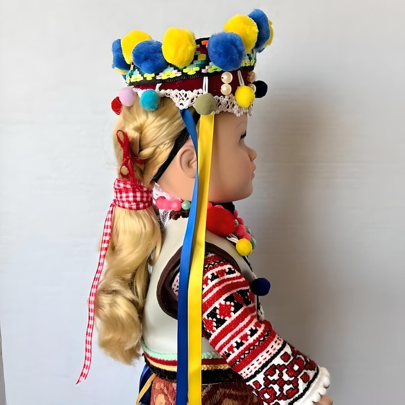 May include: A doll wearing a traditional folk costume with a colorful headpiece featuring pom-poms and ribbons. The doll has blonde hair and is wearing a red and white embroidered shirt with a white vest.