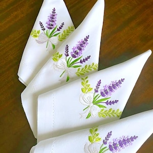 May include: Four white cloth napkins with embroidered lavender sprigs, green leaves, and a white bow. The napkins are arranged on a dark wooden surface, showcasing the detailed floral design.