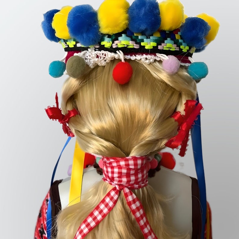May include: A doll with blonde hair styled in a braid, wearing a colorful headband with pom-poms and a red and white checkered ribbon.