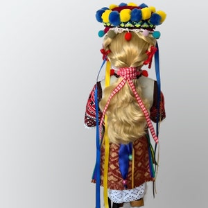 May include: A doll wearing a traditional folk costume with a colorful headpiece, red and white ribbon, and a long blonde braid.