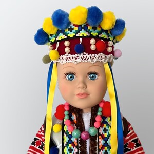 May include: A doll wearing a colorful traditional Ukrainian headdress with yellow and blue ribbons. The doll has blue eyes and is wearing a beaded necklace.