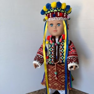 May include: A doll wearing a traditional Ukrainian folk costume with a colorful embroidered vest, a red and brown skirt, and a yellow and blue headpiece with pom-poms.