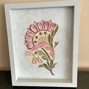 May include: Framed floral artwork featuring a stylized flower in shades of pink, cream, and olive green. The flower is set against a white background with a subtle pattern, encased in a white wooden frame. The artwork is a decorative piece.