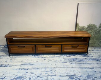 Puede incluir: Un mueble de TV de madera con tres cajones y una estantería. El mueble tiene una tapa de madera natural y una estructura de metal negro. Los cajones son de madera con diseño de lamas y tiradores negros. El mueble mide unos 183 cm de largo.